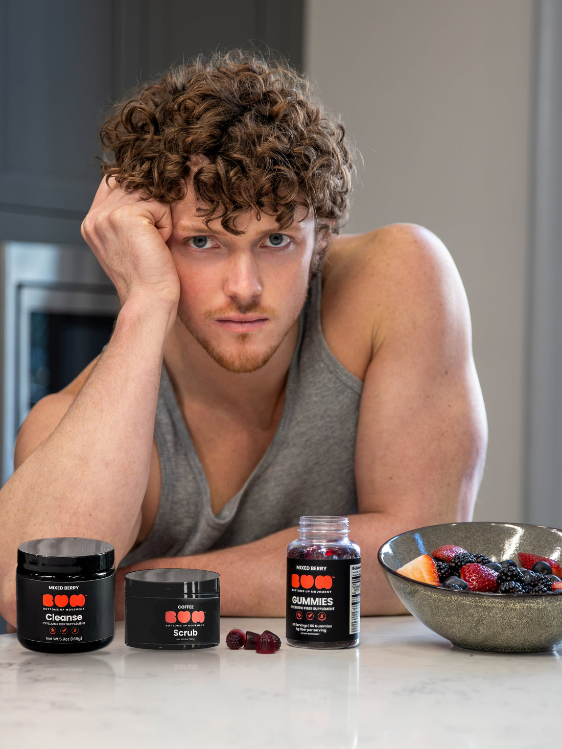 Man leaning on a kitchen counter with BUM Cleanse, Coffee Scrub, fiber gummies and a bowl of berries