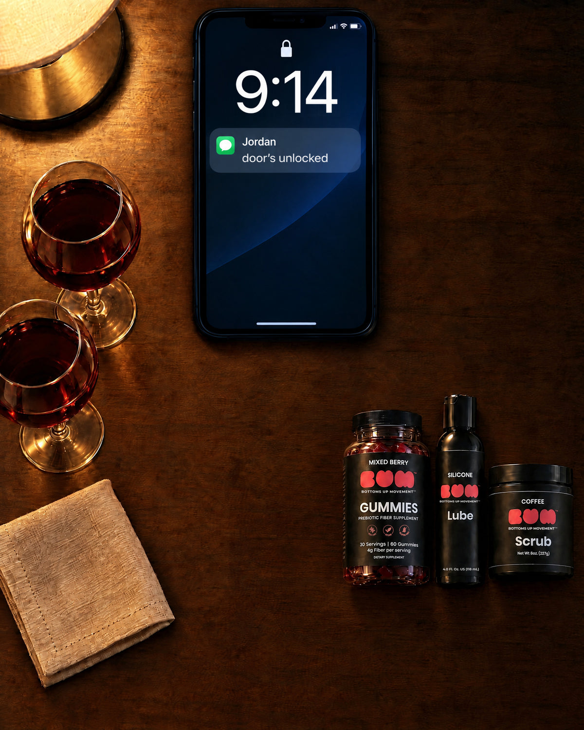 BUM Gummies, Silicone Lube and Coffee Scrub on a dark wood table next to a phone and wine glasses